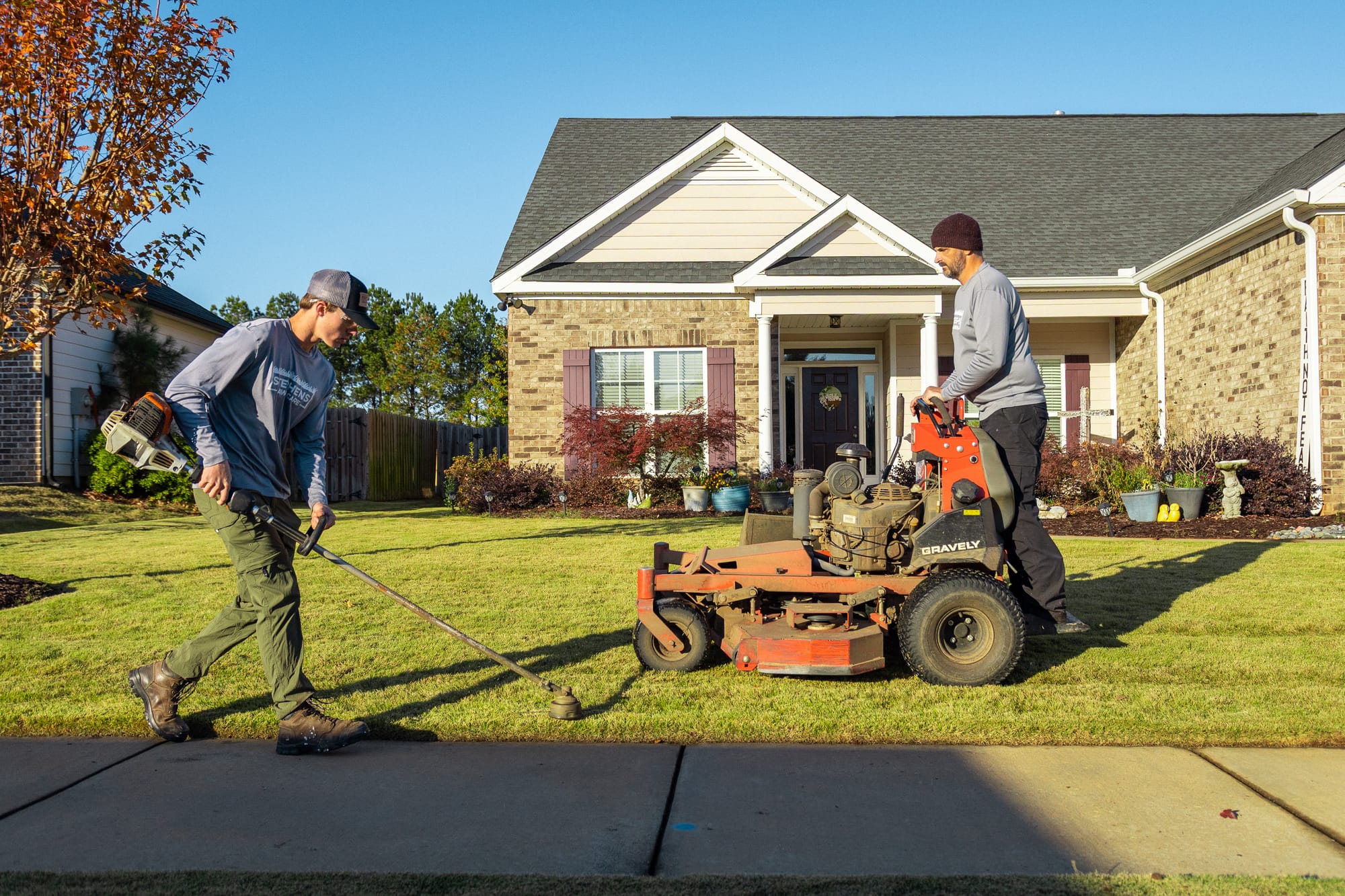 Two Stephens Lawn Care crew members trimming and mowing the same front lawn in low evening light